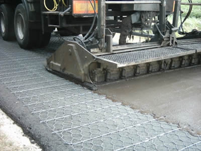 A car is extending a layer of concrete on the surface of hexagonal wire mesh, beneath the mesh is a thick layer of asphalt.