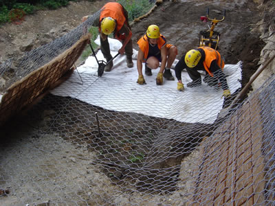 Three workers are layering green terramesh onto ground, stabilize it by reinforcing wire and rings.