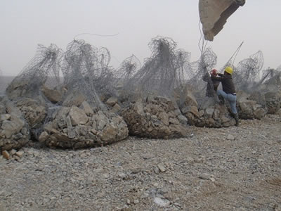 Gabion sacks are placed on a soil field, two men are hanging one of them onto a excavator.
