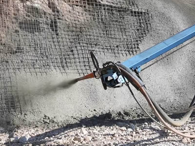 A blue cement gun is spraying concrete onto the foot of a slope.